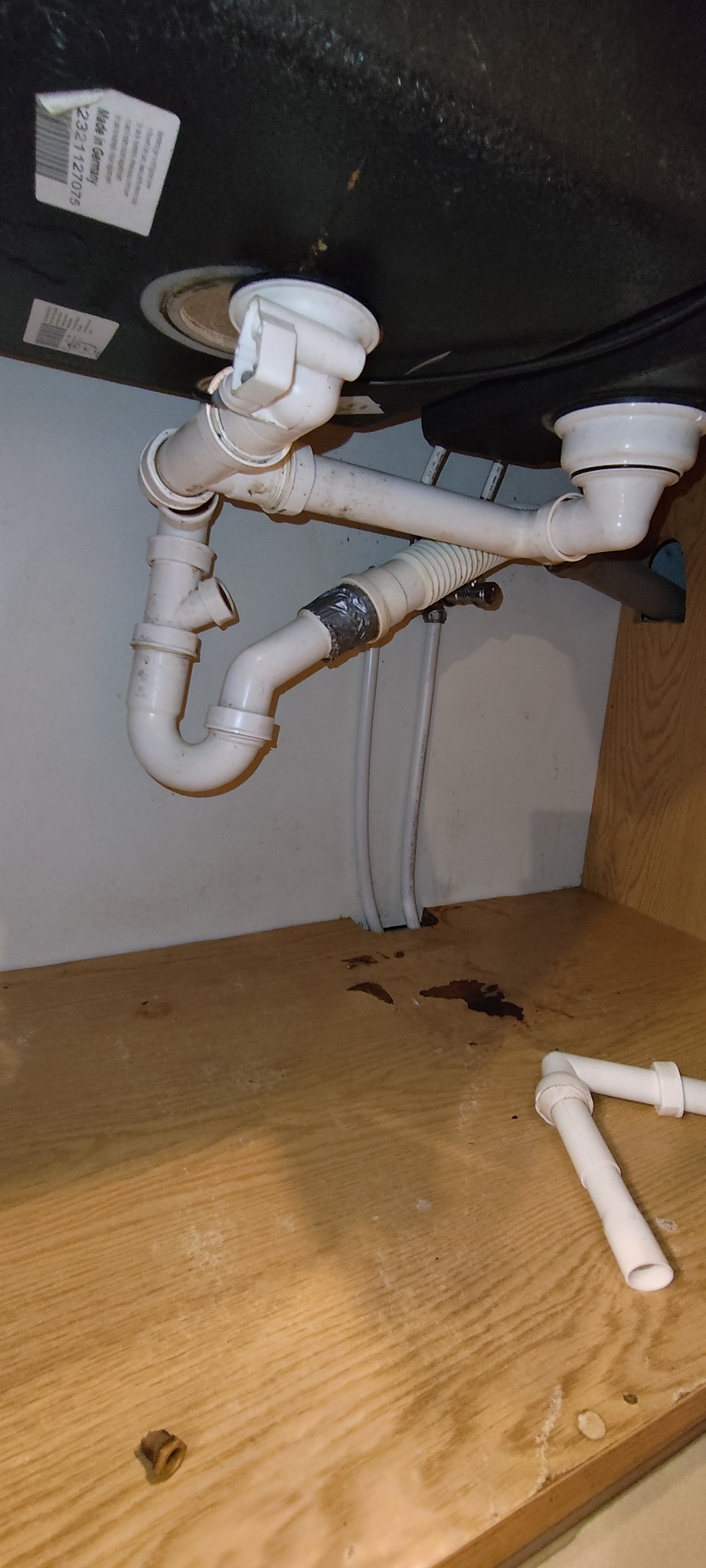 Białe rury odpływowe pod zlewem, z widocznymi śladami naprawy taśmą. Na dnie szafki plamy po wycieku i fragmenty rur. Ujęcie z dołu, w szafce pod zlewem.