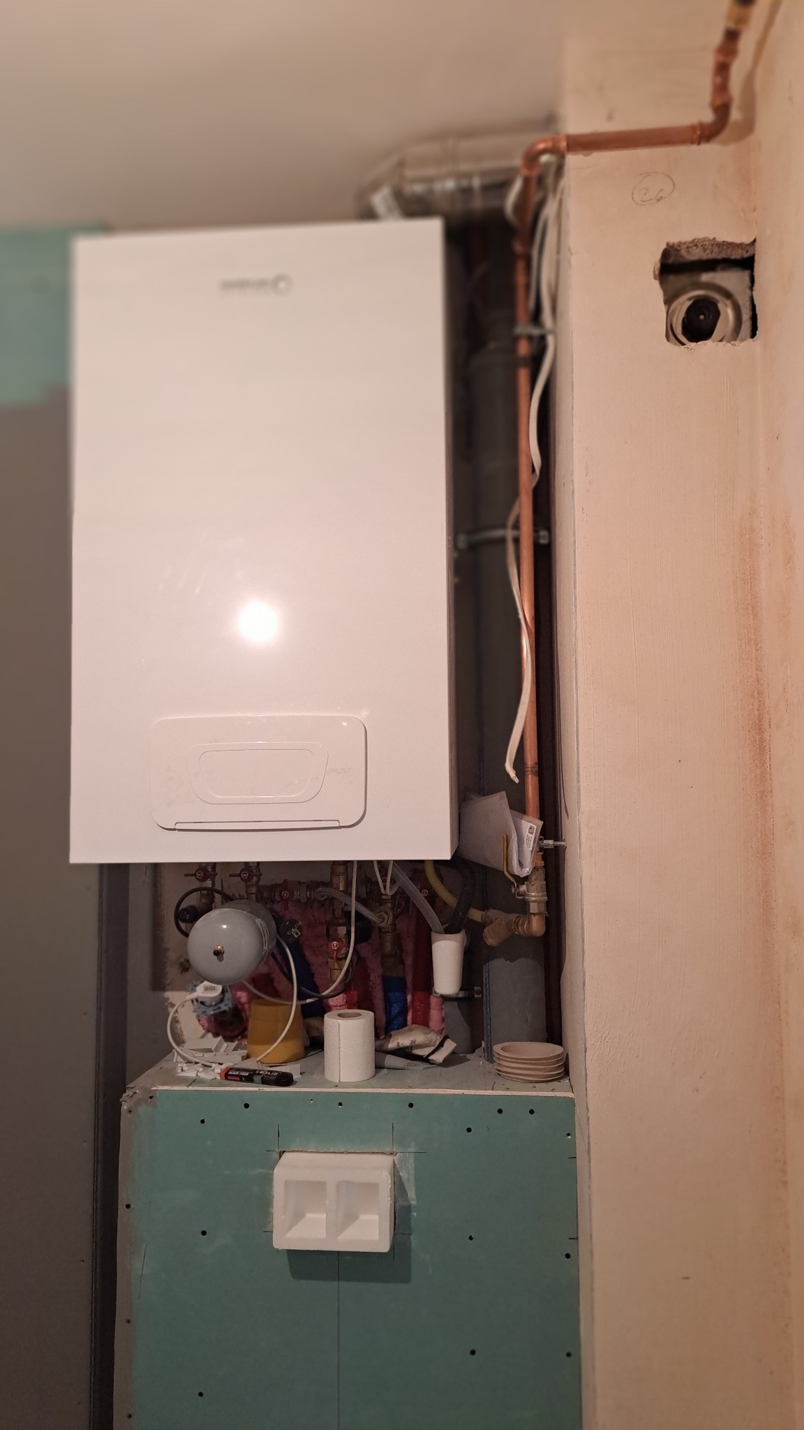 Biały piec grzewczy zamontowany na ścianie, widoczne miedziane rury instalacji, fragment ściany z otworem. Prace instalacyjne w trakcie wykończenia.