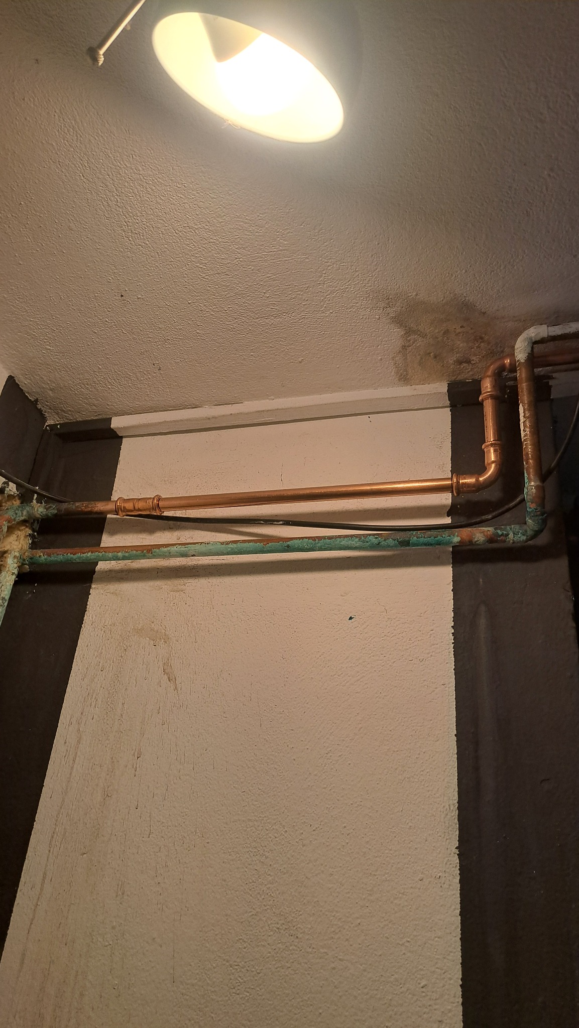 Miedziane rury instalacji wodnej, częściowo pokryte patyną, zamontowane wzdłuż ściany pod sufitem, oświetlone lampą. Widoczne złącza i fragmenty instalacji.