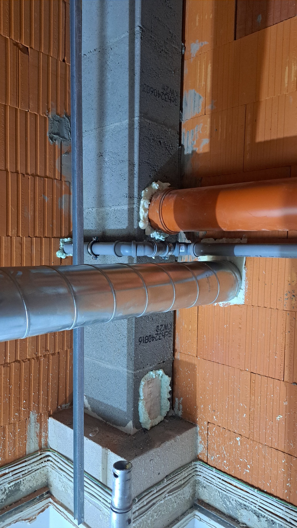 Montaż instalacji wentylacyjnej i hydraulicznej w surowym wnętrzu z ceglanymi ścianami. Widoczne rury, pianka montażowa i elementy mocujące.