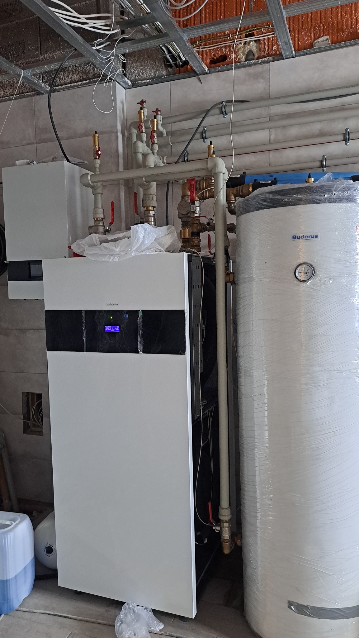 Biały piec grzewczy Buderus z podłączonymi rurami i zbiornikiem wody, widoczne elementy instalacji pod sufitem w surowym wnętrzu.