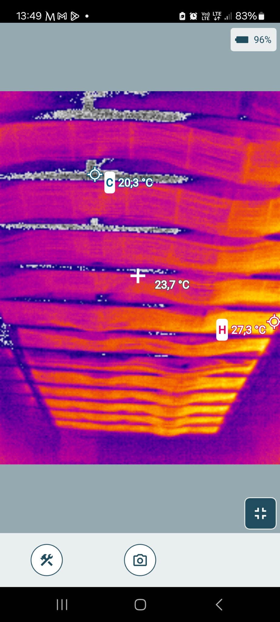 Termowizja ogrzewania podłogowego z widocznymi temperaturami rur w kolorach fioletowo-pomarańczowych. Na ekranie telefonu widoczne ikony i wskaźnik baterii.