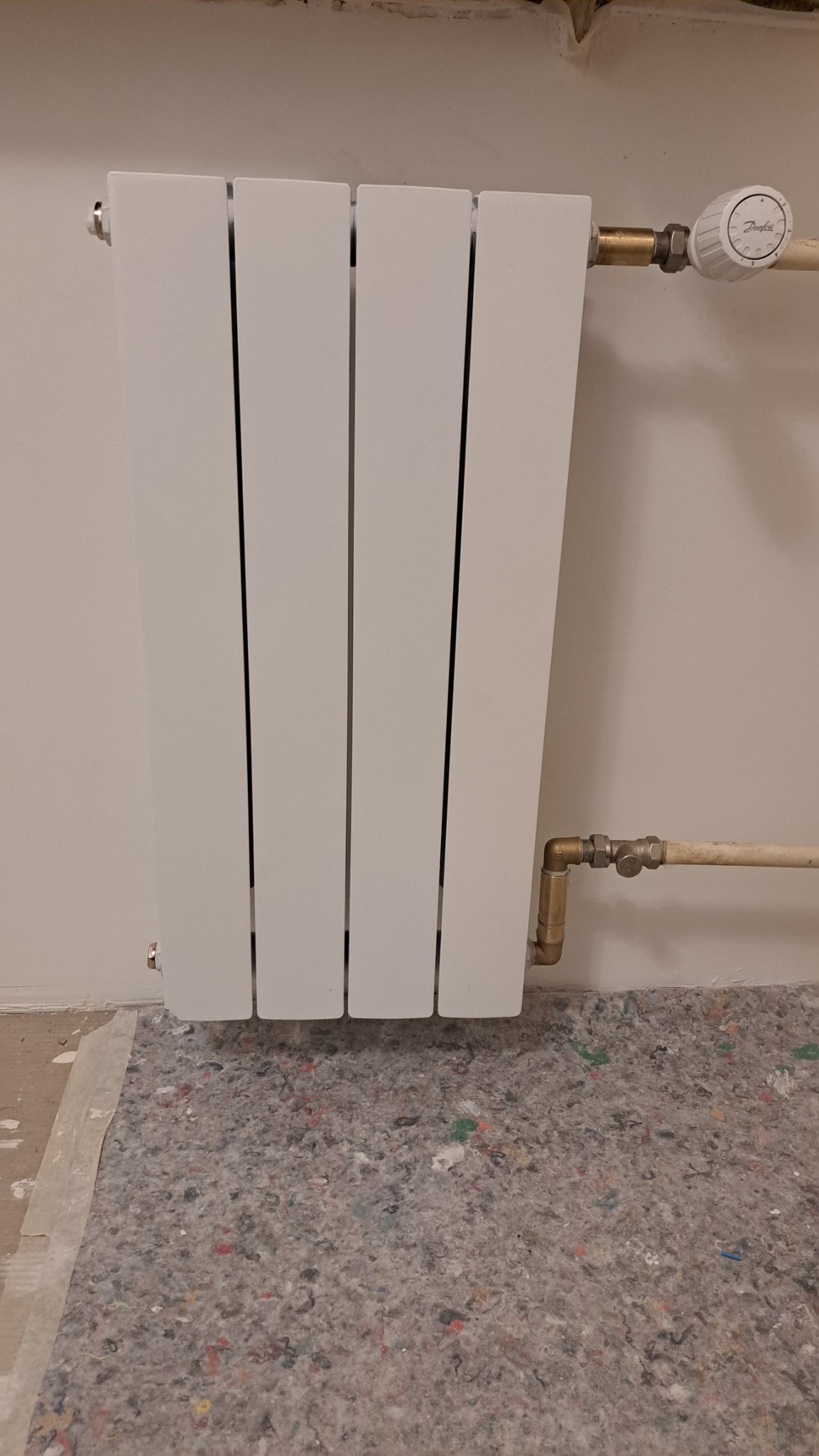 Biały, panelowy grzejnik zamontowany na ścianie, z widocznymi rurami i zaworem Danfoss. Podłoga zabezpieczona szarą matą ochronną.