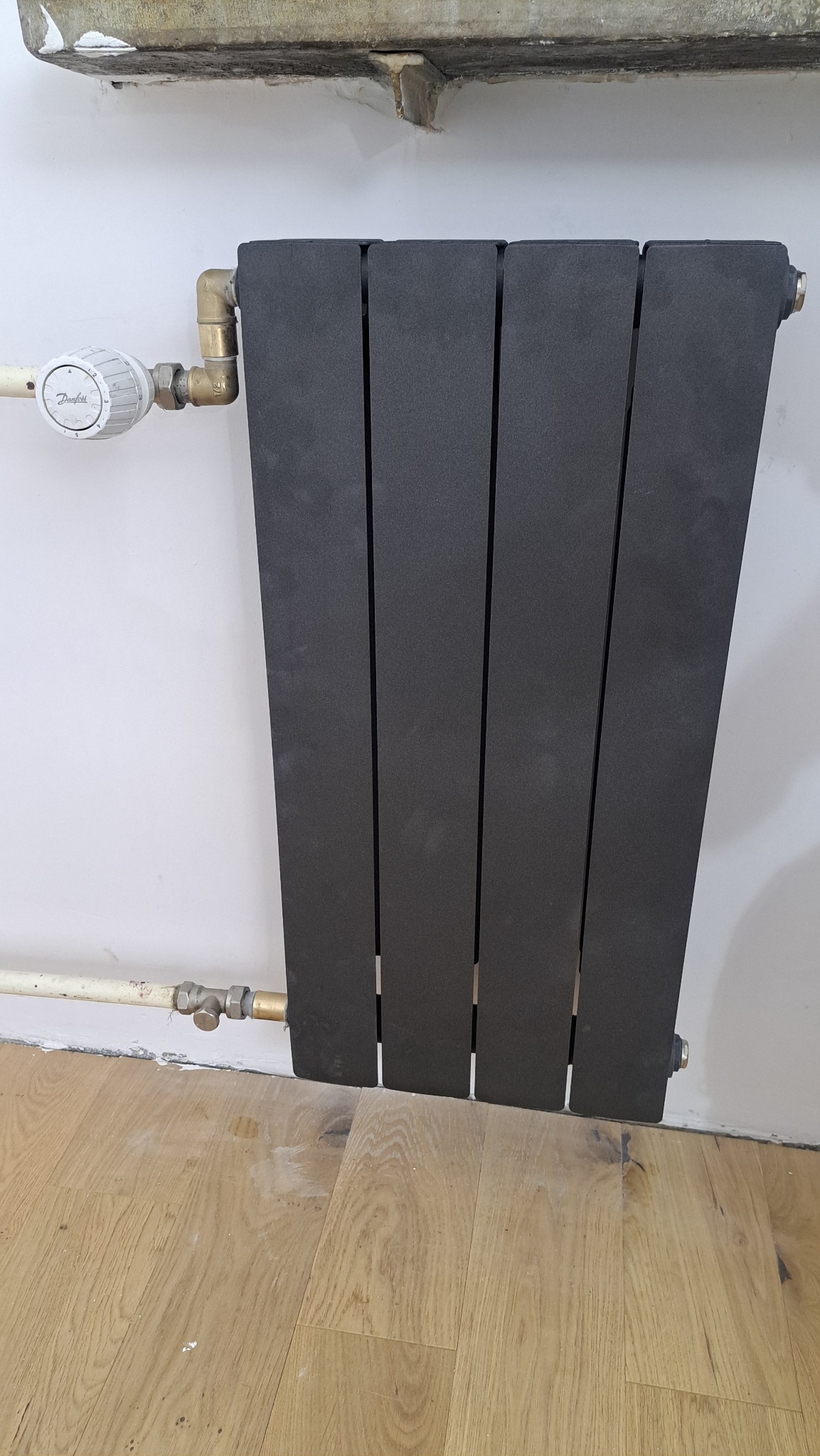 Czarny, panelowy grzejnik zamontowany na białej ścianie, z widocznymi rurami i zaworem termostatycznym. Podłoga drewniana. Ujęcie z bliska, detale instalacji grzewczej.