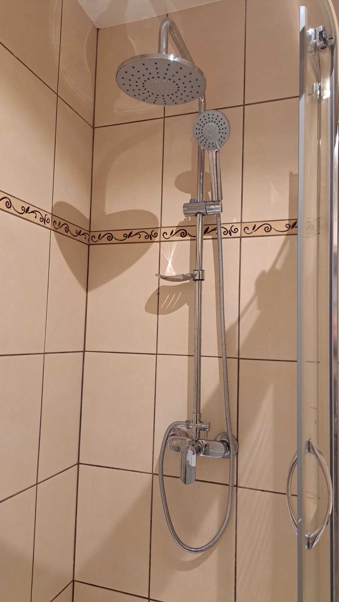 Bateria prysznicowa z deszczownicą i słuchawką, zamontowana na beżowych płytkach z dekoracyjnym paskiem. Widoczny fragment kabiny prysznicowej.