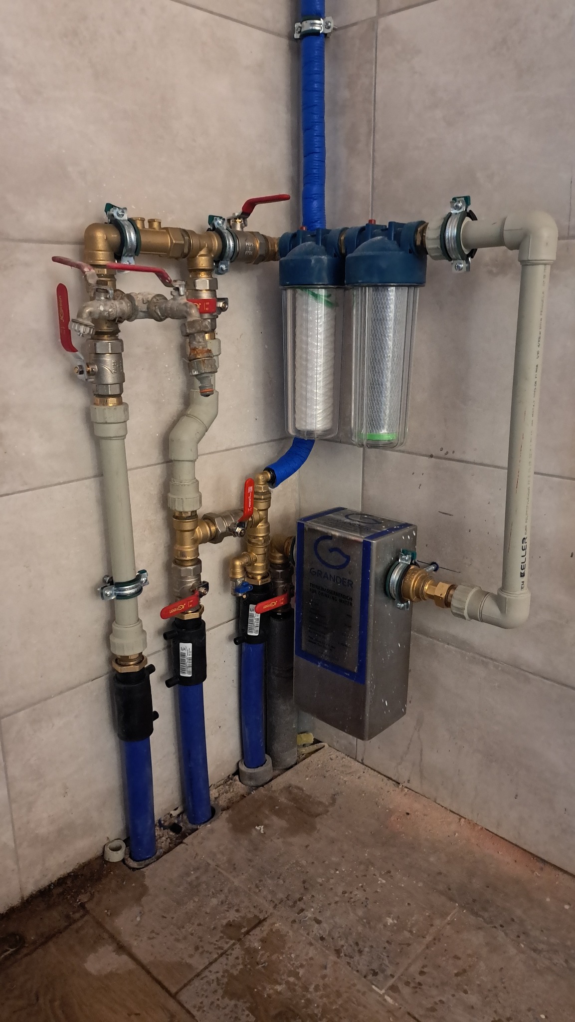 Kompleksowa instalacja hydrauliczna z filtrami wody i zaworami, rury miedziane i plastikowe, widoczne łączenia i mocowania, schludne wykonanie przy ścianie z płytek.