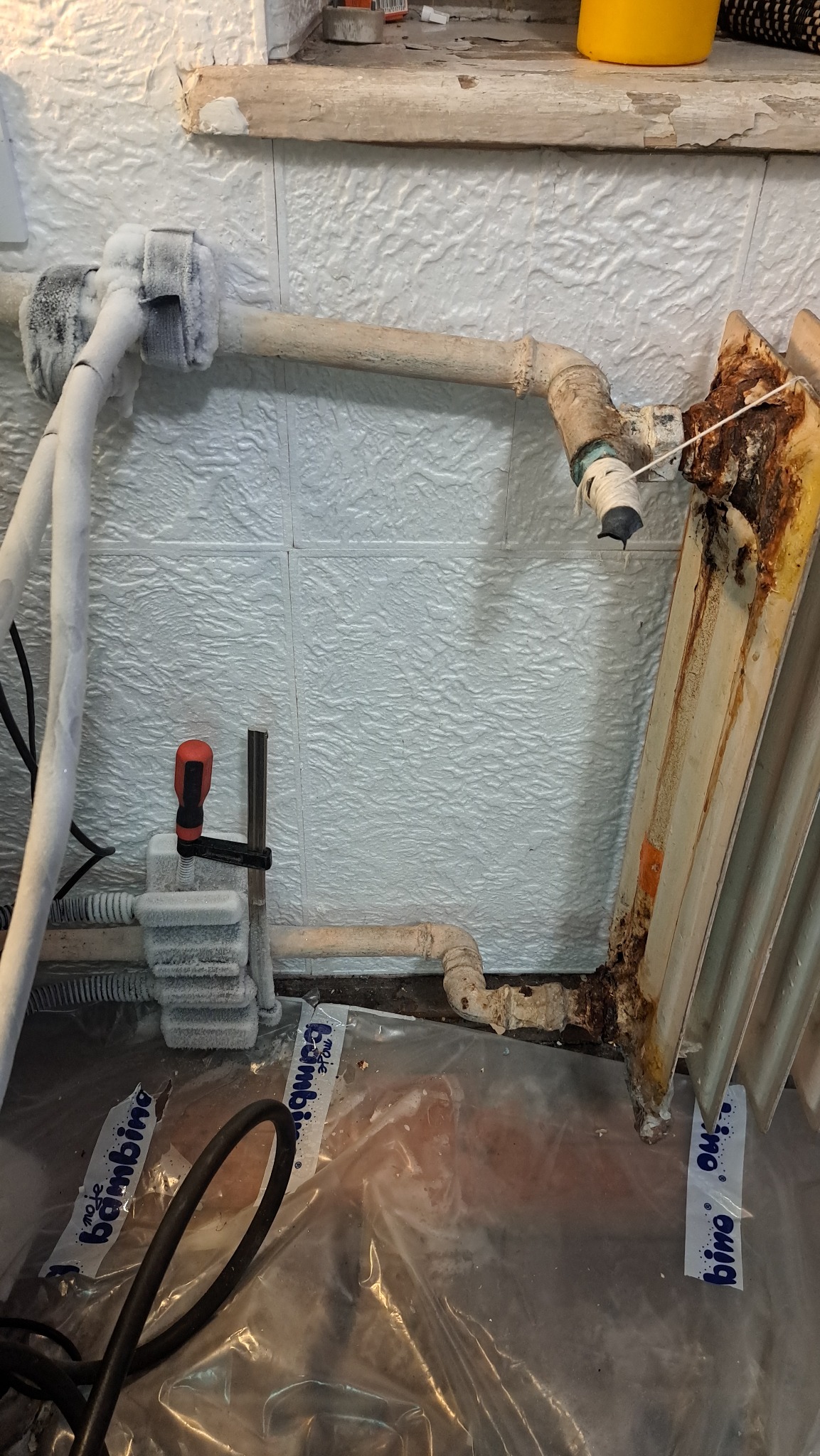 Zamrożone rury i skorodowany grzejnik w trakcie naprawy, zabezpieczone folią. Widoczne narzędzia hydrauliczne i biała, teksturowana ściana w tle.