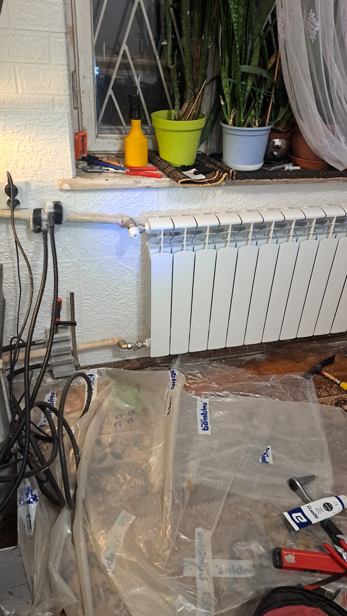 Biały grzejnik panelowy zamontowany pod oknem, widoczne rury doprowadzające, narzędzia i folia ochronna na podłodze. Świeżo po montażu, w tle rośliny doniczkowe.