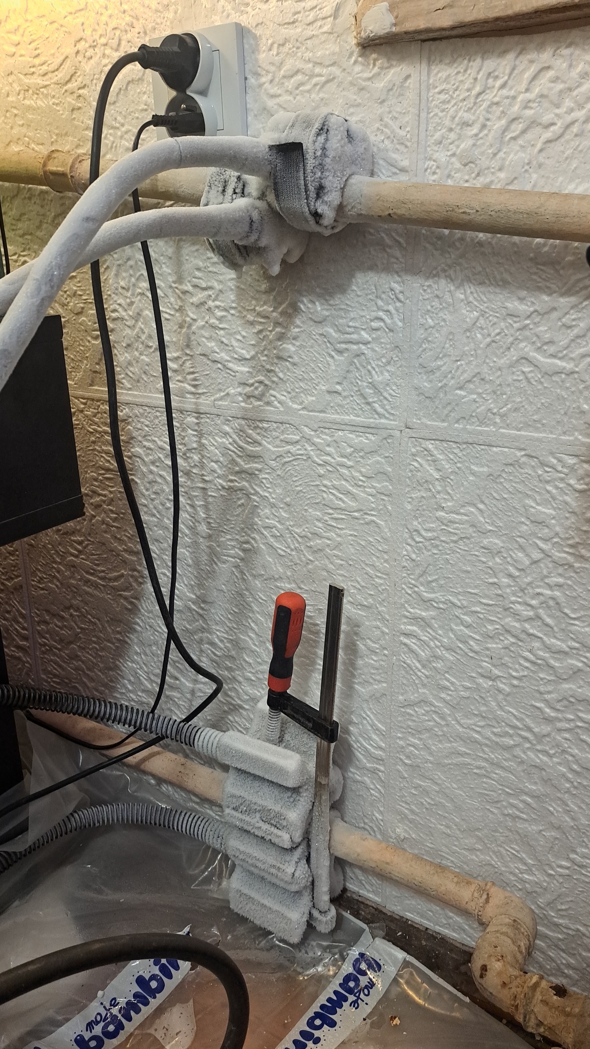 Zamrożone rury instalacji wodnej zabezpieczone zaciskiem śrubowym, widoczne narzędzia i gniazdko elektryczne na tle białej ściany z fakturą.