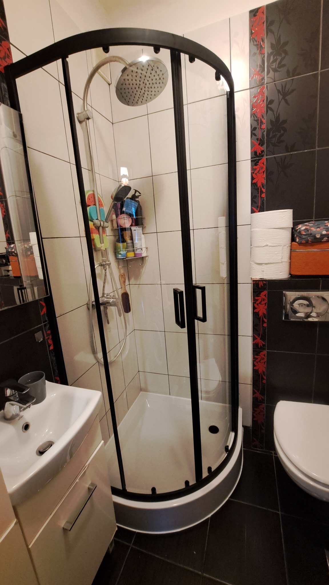 Nowoczesna kabina prysznicowa z czarnymi profilami w wykafelkowanej łazience. Widoczna armatura i półka z kosmetykami. Uwagę zwracają czarno-białe płytki z czerwonym akcentem.