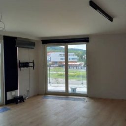 Vasyl Hapiyk - Jasne wnętrze pokoju z panelami podłogowymi, widocznym klimatyzatorem, zamontowanym uchwytem na telewizor i oknem balkonowym z widokiem na budynek mieszkalny.