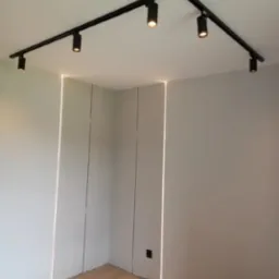 Nowoczesne oświetlenie sufitowe z czarnymi reflektorami na szynie, uzupełnione pionowymi liniami świetlnymi na ścianach w minimalistycznym wnętrzu.