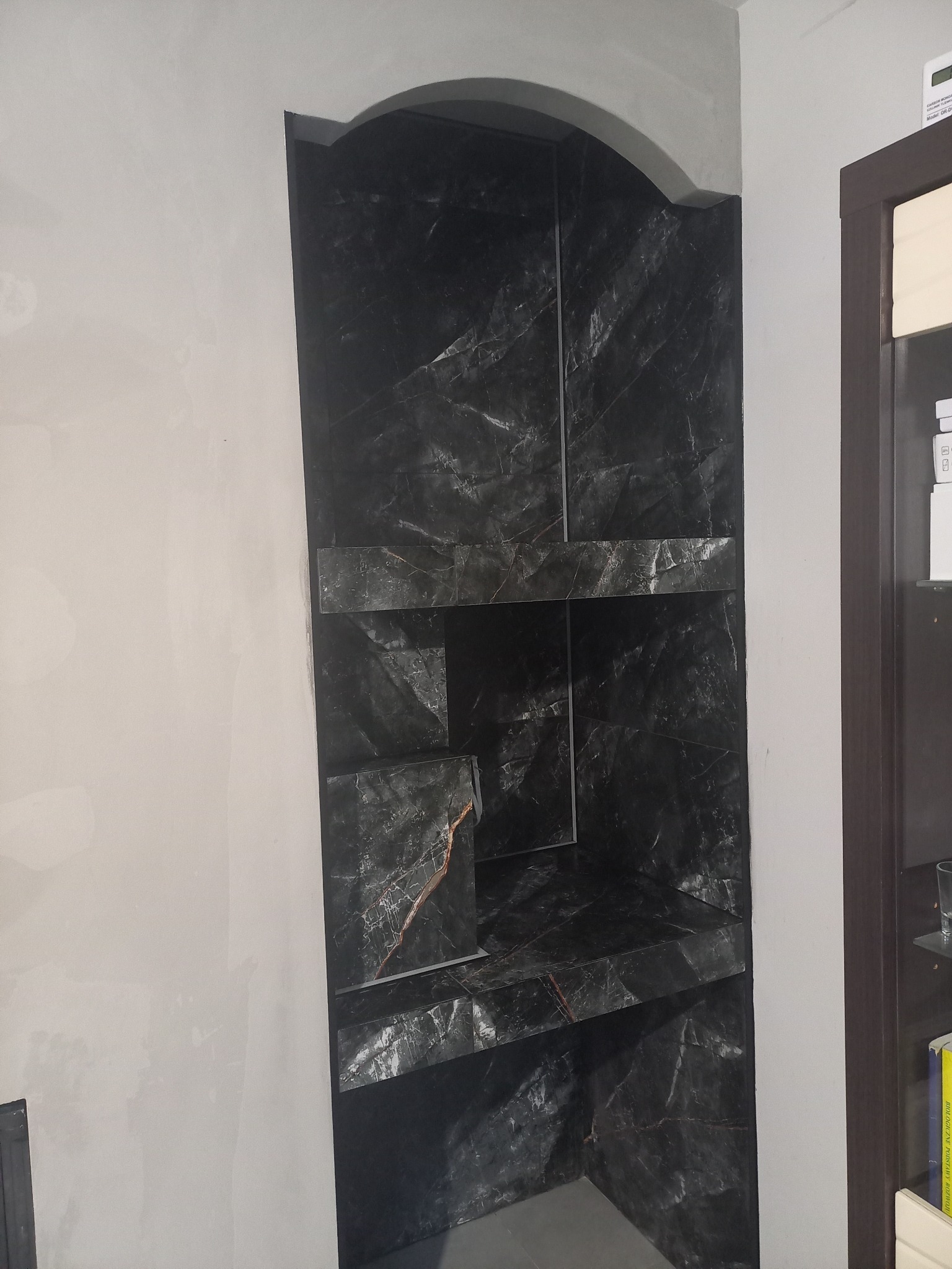 Narożna wnęka z półkami wykończona czarnym marmurem z pomarańczowymi żyłkami, na tle szarej ściany, widoczny fragment futryny drzwi.
