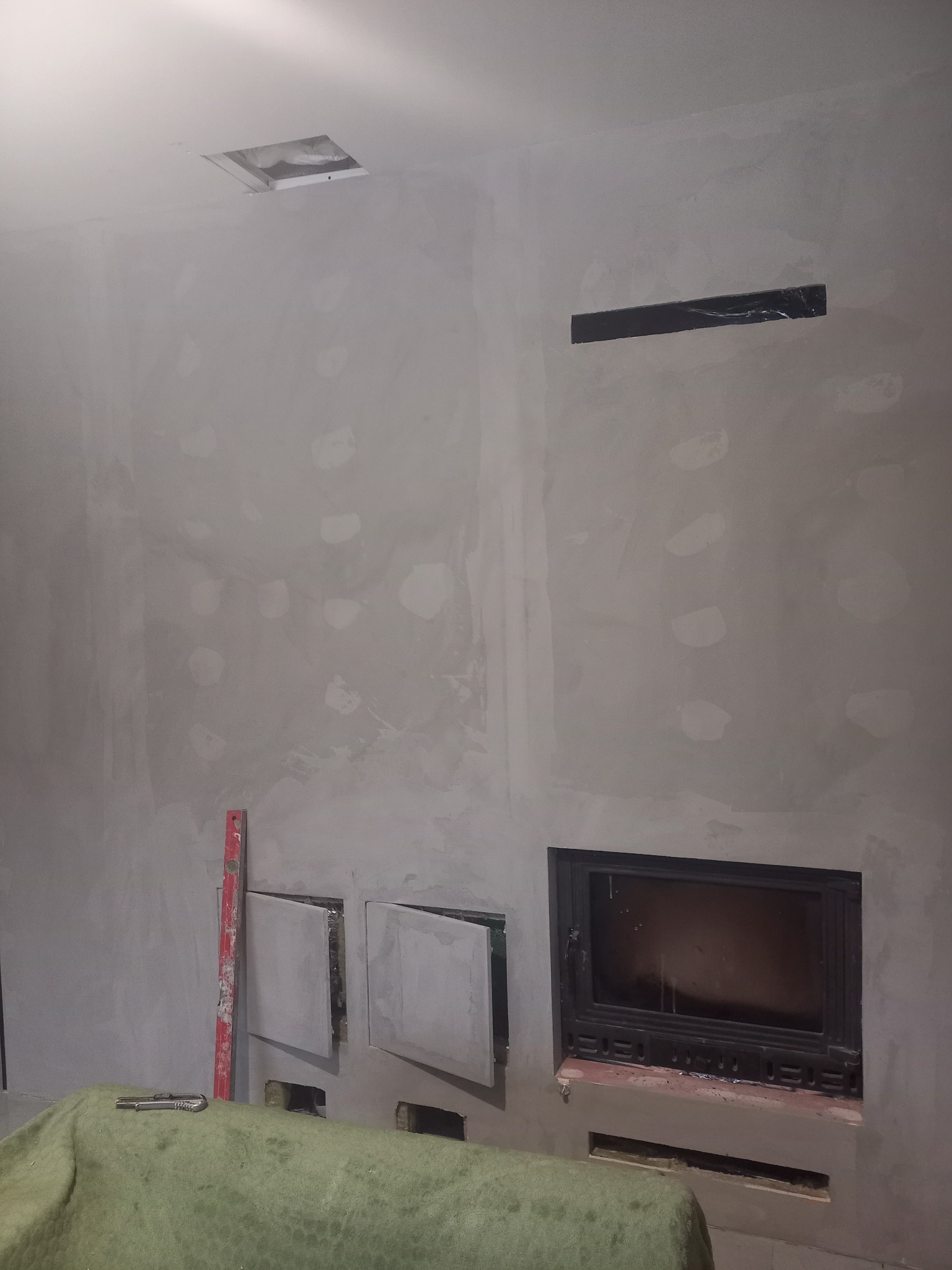 Ściana w trakcie szpachlowania z wnęką na kominek, widoczne otwory rewizyjne, poziomica i nóż na zielonym materiale zabezpieczającym podłogę.