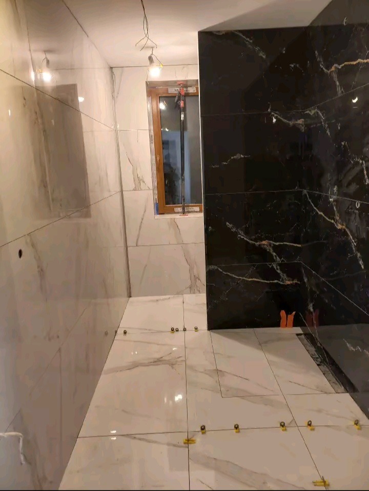 Łazienka w trakcie wykańczania: białe i czarne marmurowe płytki na ścianach i podłodze, żółte kliny poziomujące płytki podłogowe, okno z widokiem na zewnątrz.