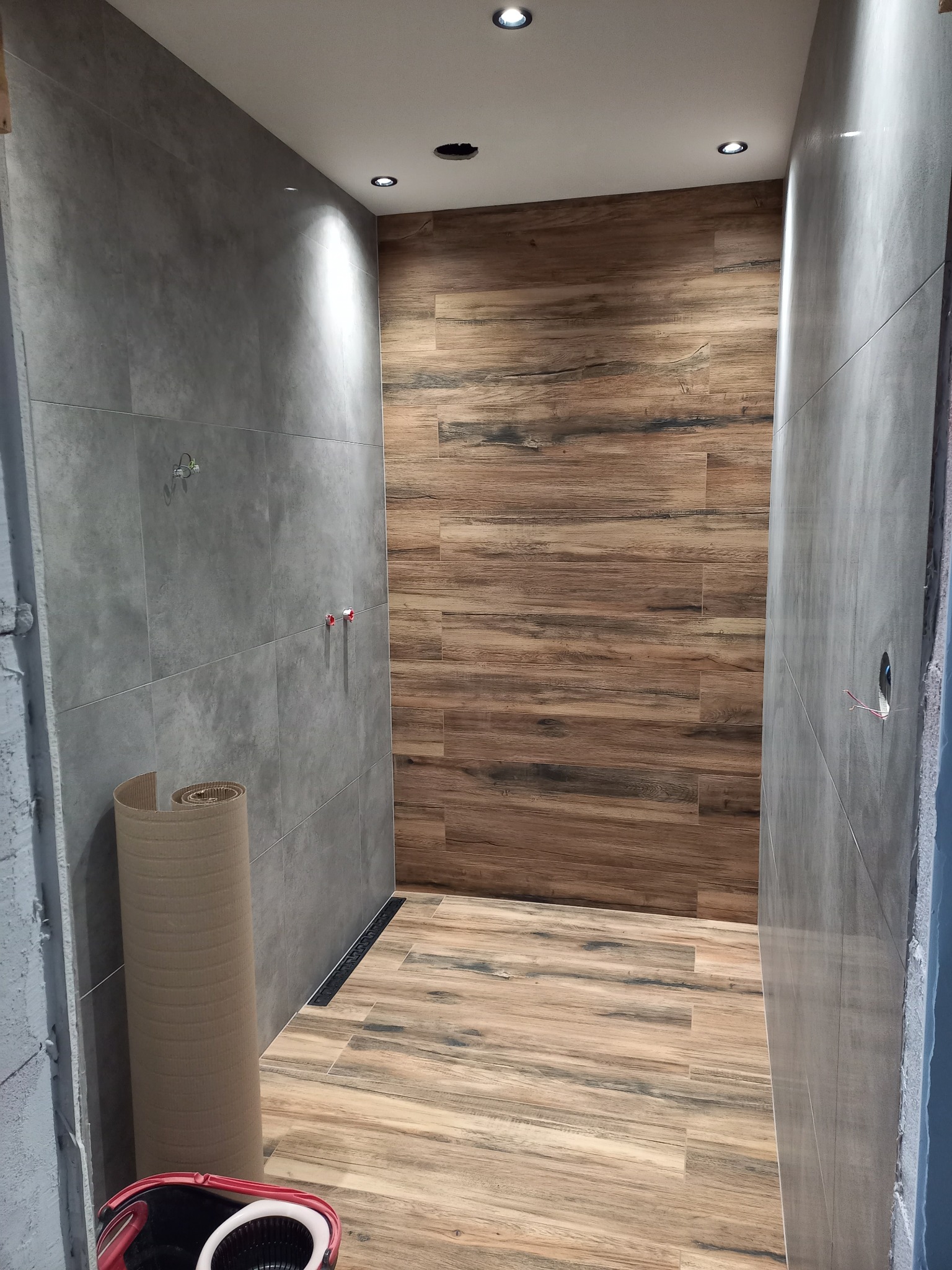 Wnętrze łazienki w trakcie prac, z szarymi płytkami na ścianach, drewnopodobną okładziną na jednej ze ścian i podłodze, widoczny odpływ liniowy, rolka zabezpieczająca i niedokończone instalacje.