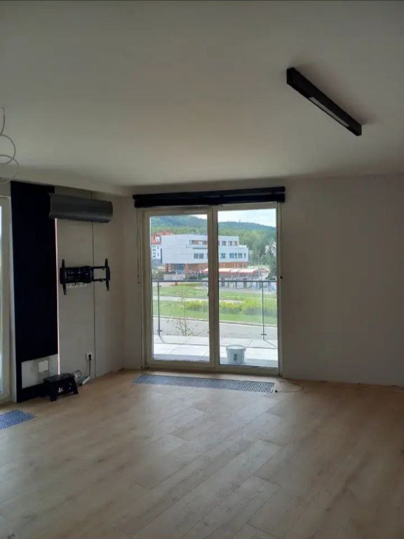 Jasne wnętrze pokoju z panelami podłogowymi, widocznym klimatyzatorem, zamontowanym uchwytem na telewizor i oknem balkonowym z widokiem na budynek mieszkalny.