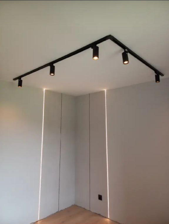 Nowoczesne oświetlenie sufitowe z czarnymi reflektorami na szynie, podkreślone pionowymi liniami świetlnymi w ścianie, drewniana podłoga.