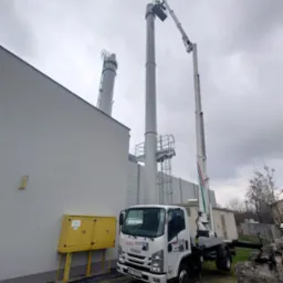 Pracownik w koszu podnośnika teleskopowego przy kominie przemysłowym, biała ciężarówka Isuzu na tle budynku z żółtą skrzynką gazową, pochmurne niebo.