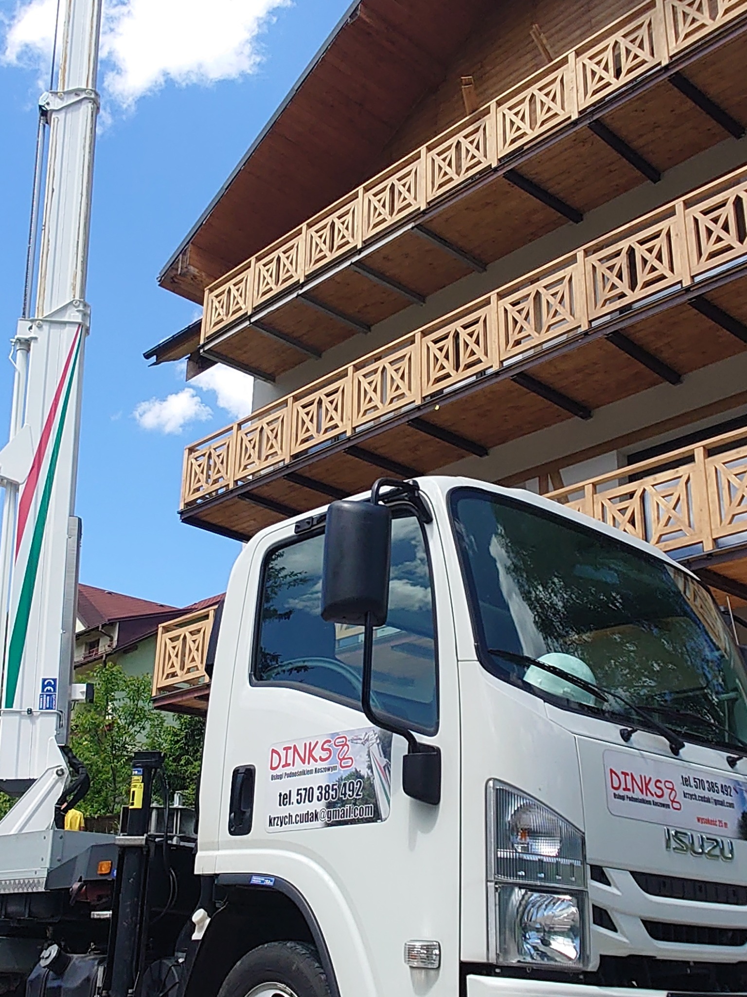 Biały samochód z podnośnikiem koszowym firmy Dinks Usługi Podnośnikiem Koszowym na tle budynku z drewnianymi balkonami i błękitnym niebem.