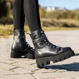 Czarne, skórzane botki na grubej podeszwie, ozdobione ćwiekami, na tle chodnika i zieleni. Widoczne czarne rajstopy i fragment spódnicy.
