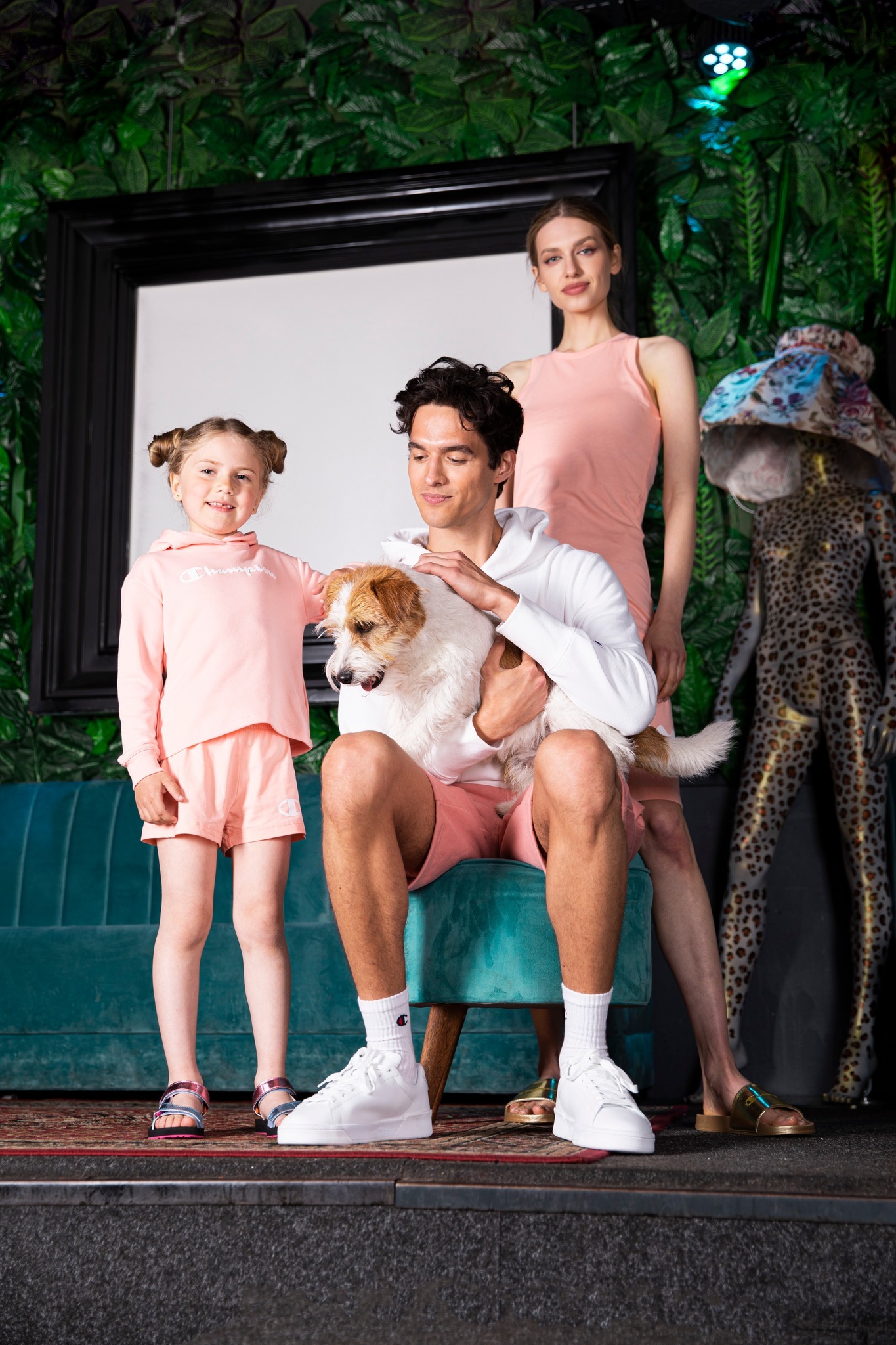 Stylizowana sesja rodzinna: ojciec z psem, córka w różowym dresie Champion i matka w sukience, na tle tropikalnej scenografii i turkusowej sofy.