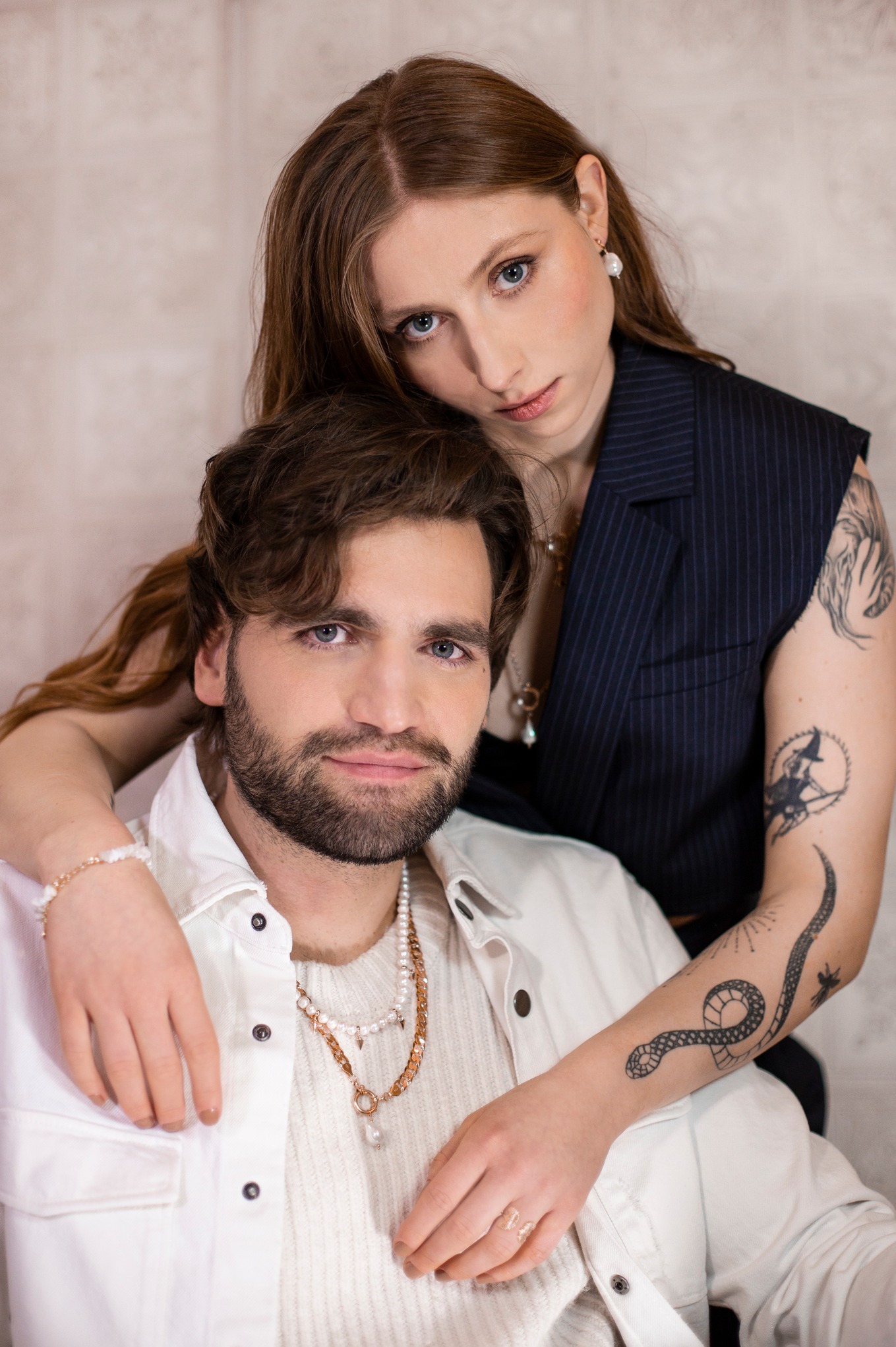 Portret pary: mężczyzna z brodą w białej kurtce i kobieta z tatuażami w kamizelce, oboje z biżuterią, na jasnym tle. Sesja wizerunkowa.