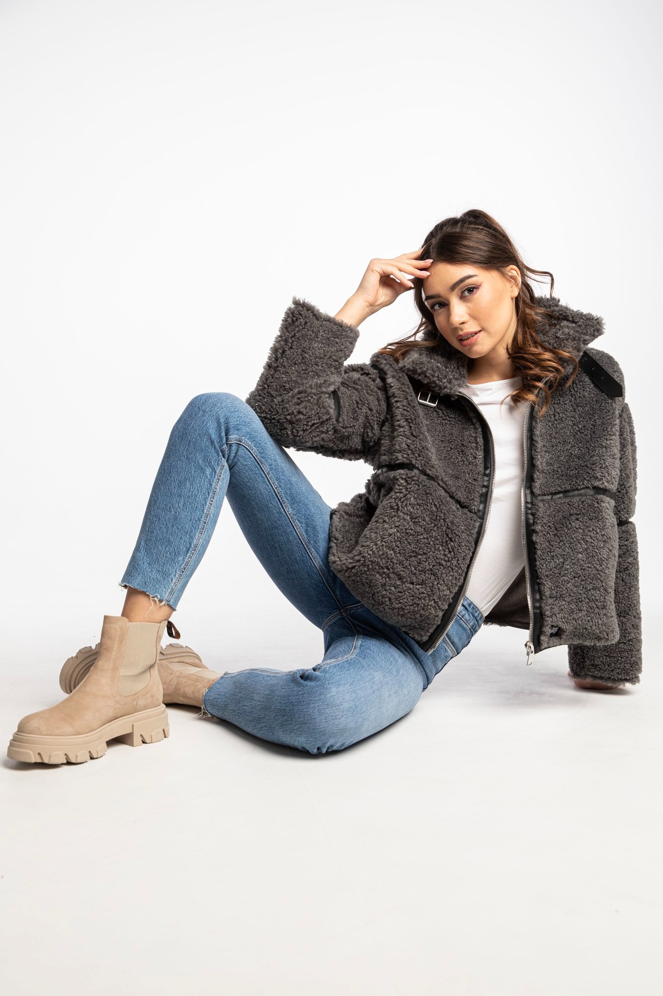 Kobieta w jeansach i kurtce z baranka, siedząca na białym tle, prezentuje modny look z beżowymi botkami na platformie.