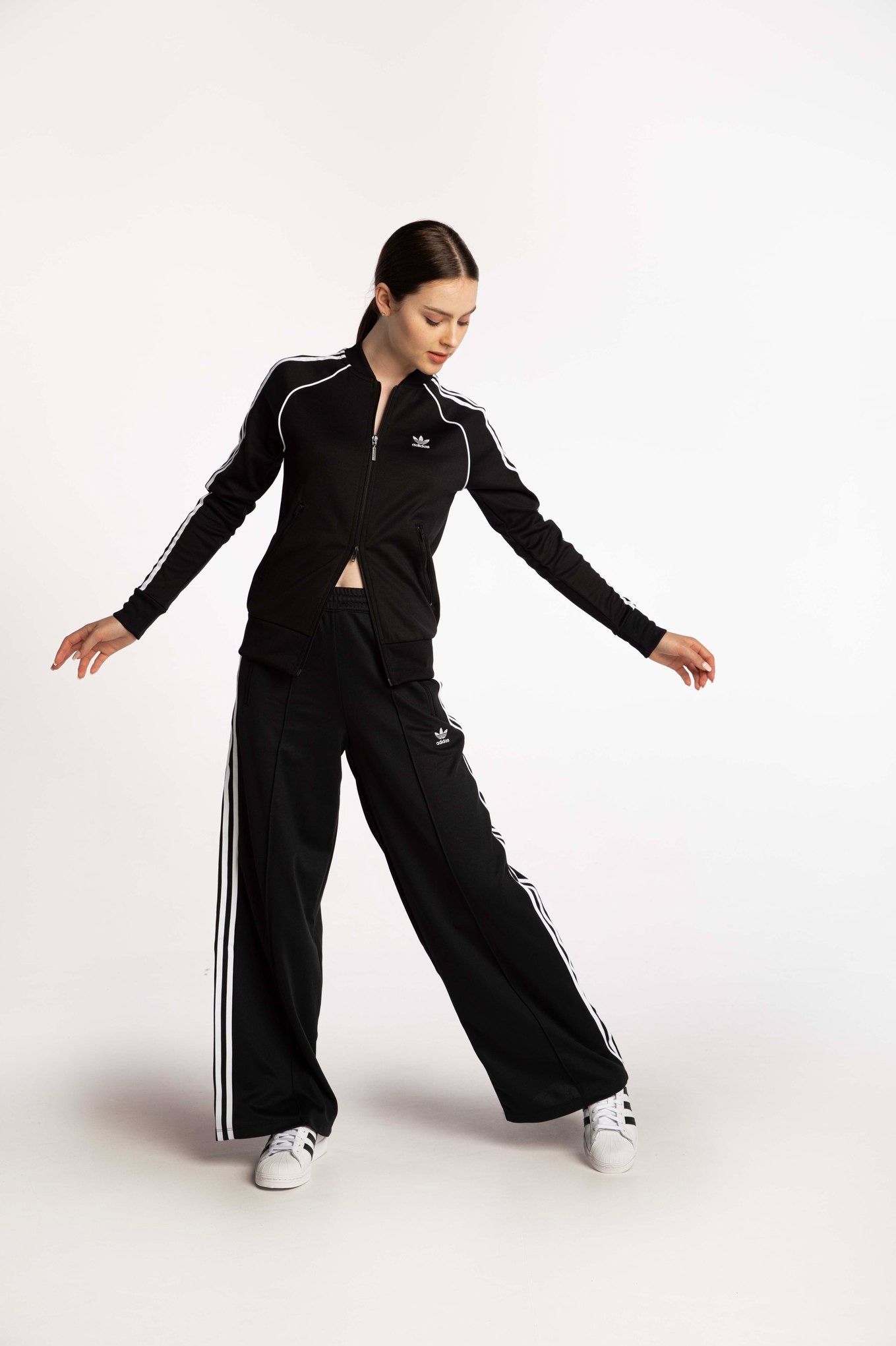 Modelka w czarnym dresie Adidas z białymi paskami, pozująca na białym tle. Uwagę zwracają szerokie spodnie i delikatny makijaż.