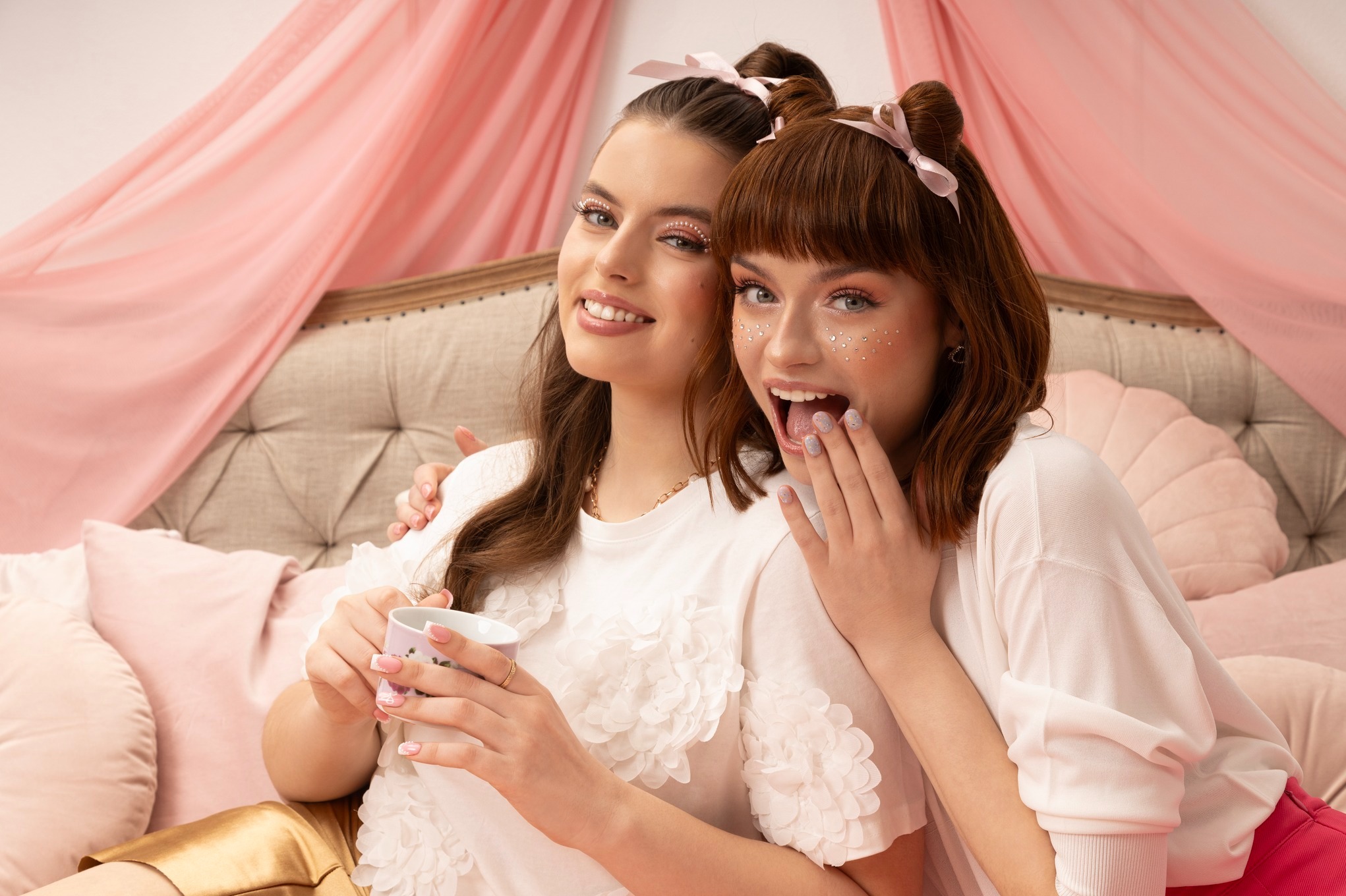 Dwie uśmiechnięte dziewczyny z brokatowym makijażem, siedzące na łóżku z baldachimem, jedna trzyma filiżankę. Romantyczna sesja w pastelowych kolorach.