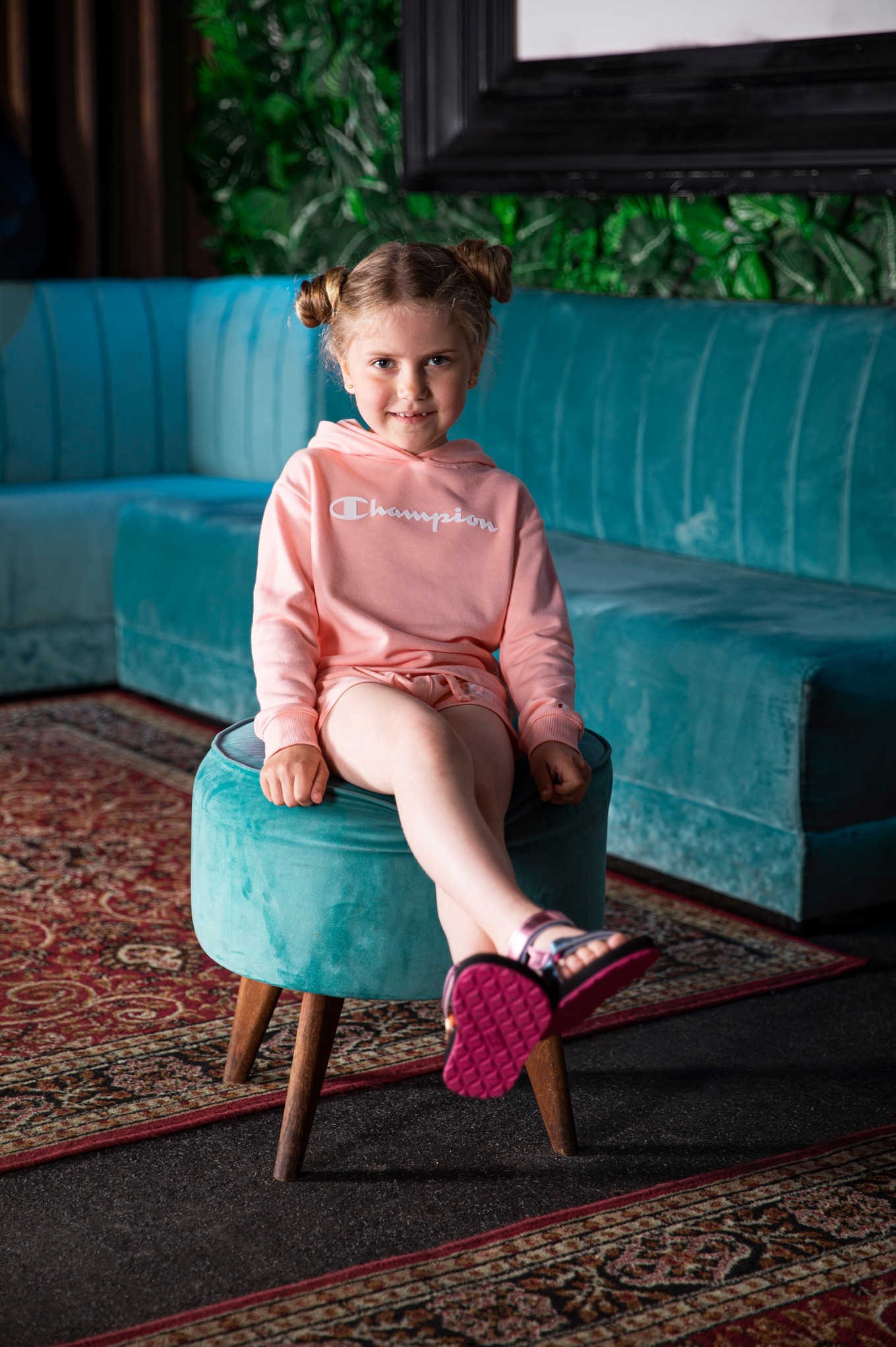 Uśmiechnięta dziewczynka w różowym dresie siedzi na turkusowym pufie, na tle sofy w tym samym kolorze i zielonej ściany. Urocza fryzura z dwoma koczkami.