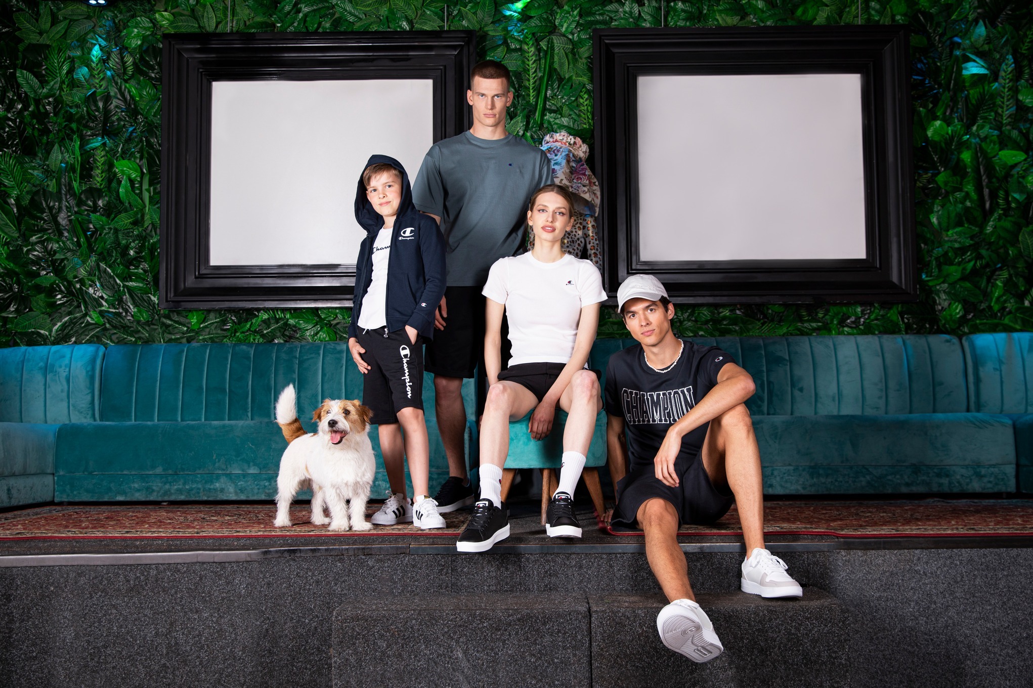 Rodzinna sesja zdjęciowa w studiu z zielonym tłem i ramami. Cztery osoby i pies pozują w sportowych ubraniach marki Champion. Nietypowa aranżacja wnętrza.