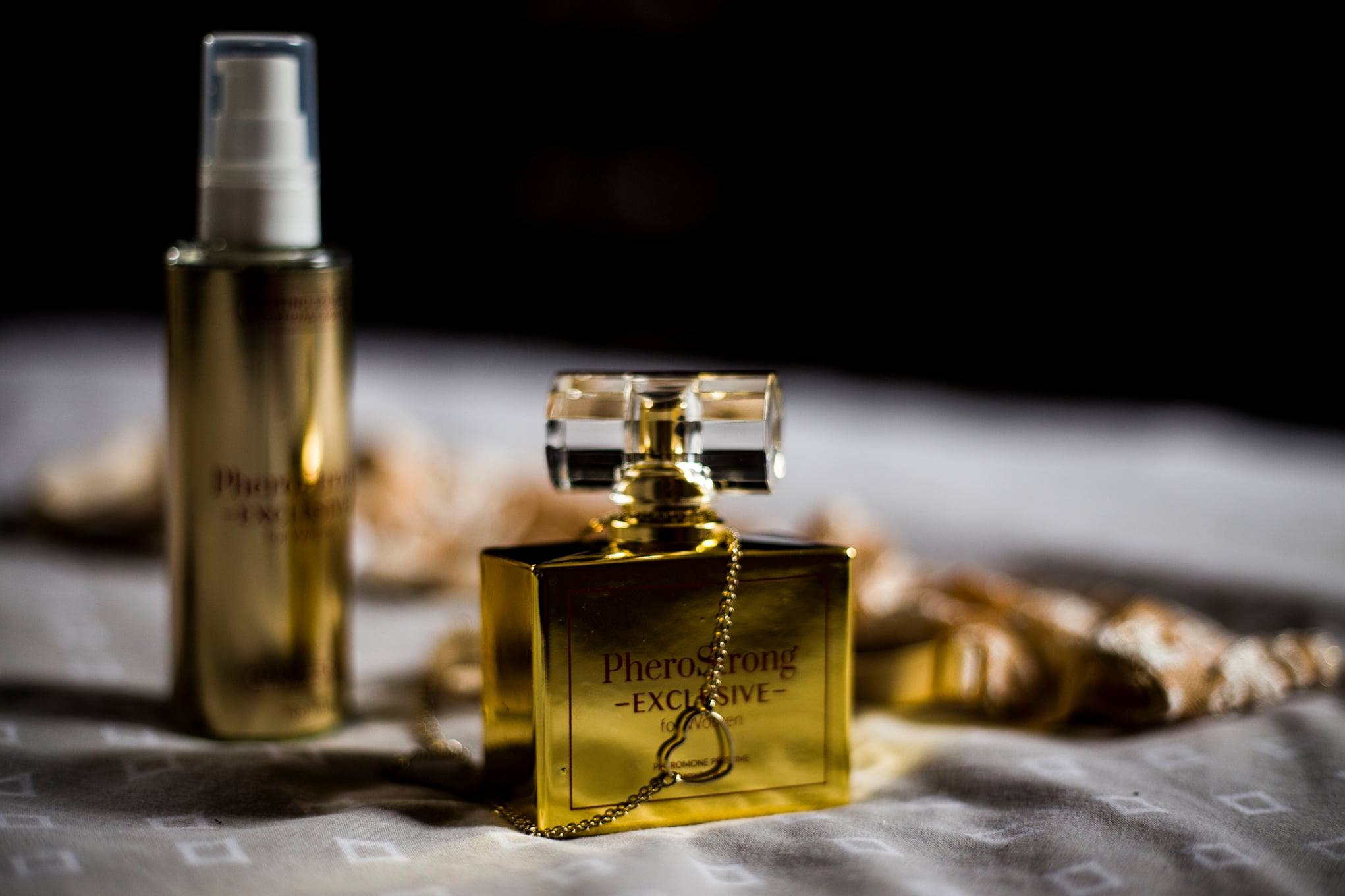 Złoty flakon perfum PheroStrong Exclusive z łańcuszkiem w kształcie serca, obok butelka z atomizerem, na wzorzystej tkaninie w przyciemnionym otoczeniu.