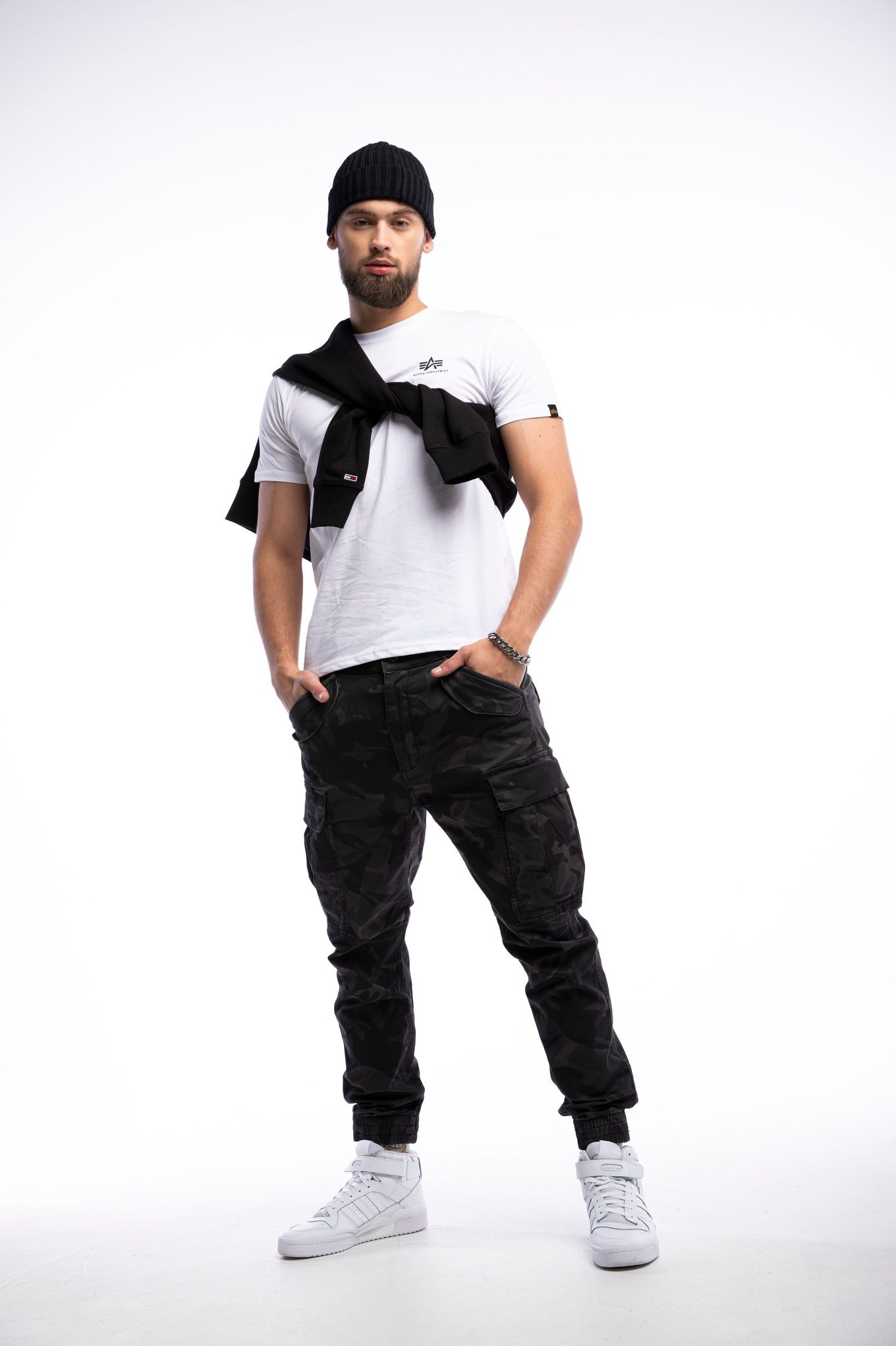 Mężczyzna w białej koszulce, czarnych spodniach cargo i czapce, pozuje na białym tle. Na ramionach ma przewiązaną czarną bluzę. Całość w stylu fashion.