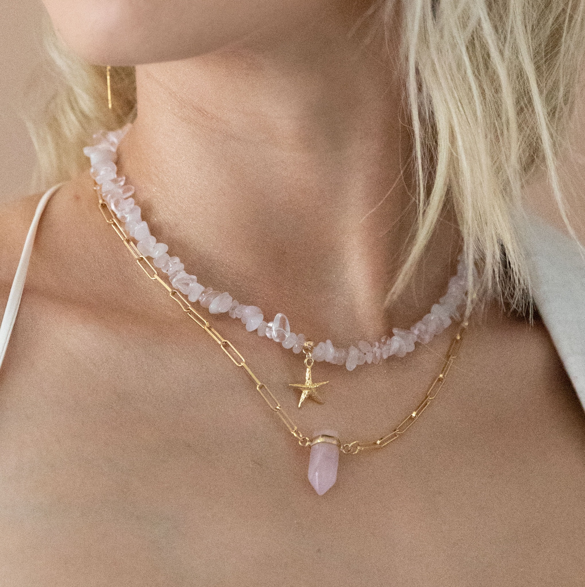 Kobieca szyja z dwoma naszyjnikami: z kamieni rózowych i złotym łańcuszkiem. Na jednym wisi gwiazdka, na drugim kryształ. Subtelne oświetlenie, kremowe tło.