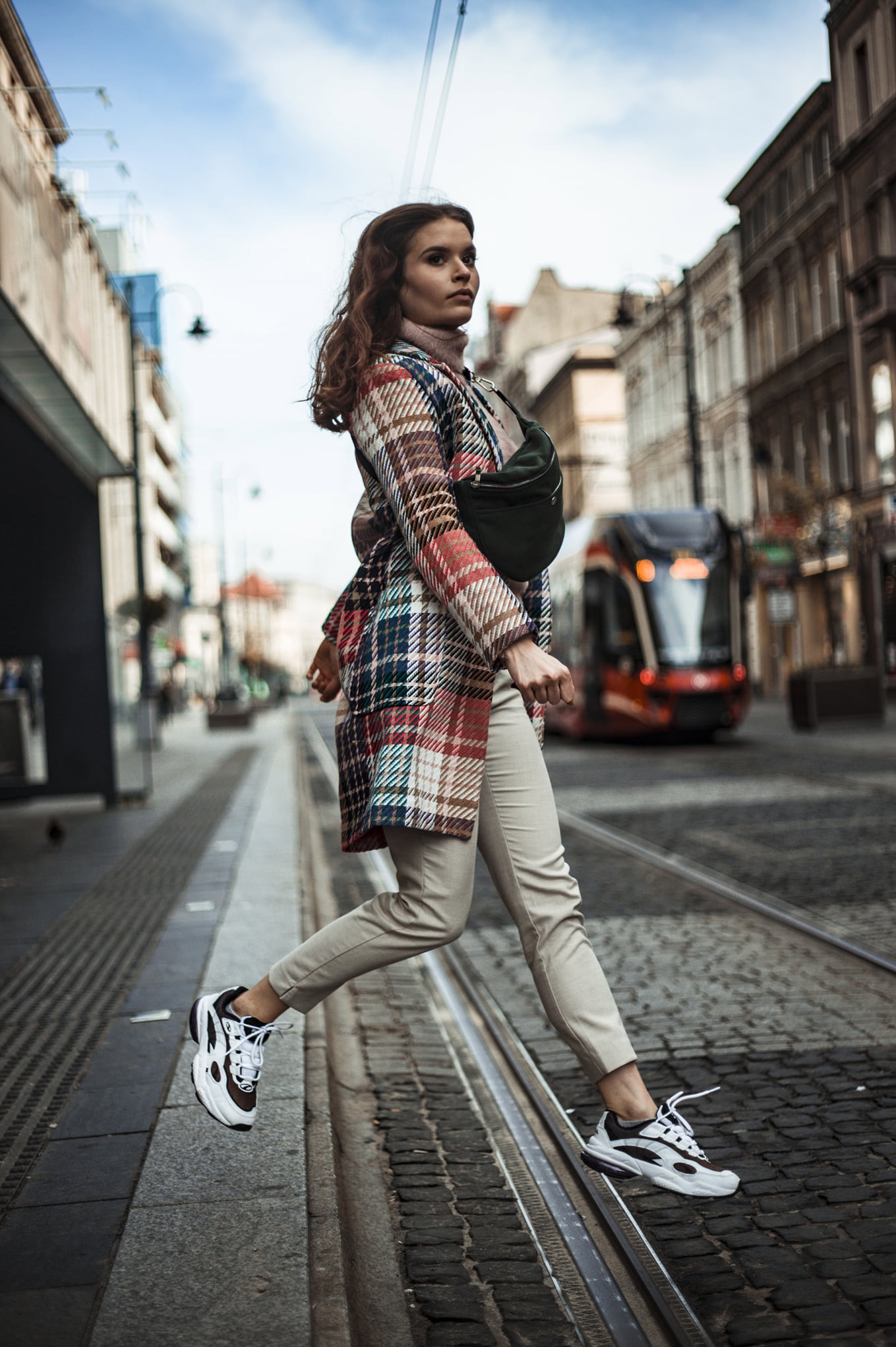 Kobieta w płaszczu w kratę skacze przez torowisko tramwajowe na tle miejskiej zabudowy i nadjeżdżającego tramwaju. Dynamiczne ujęcie z niskiej perspektywy.