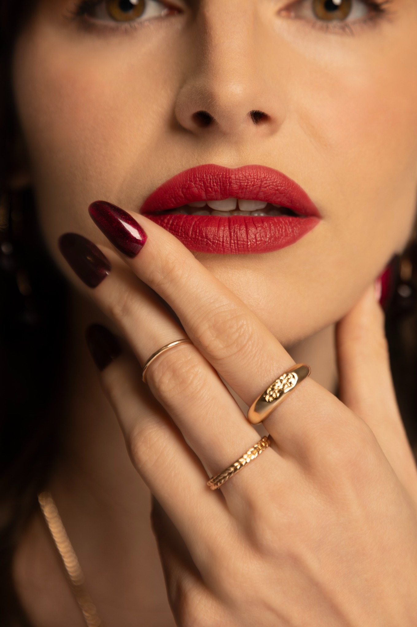 Zbliżenie na usta kobiety w czerwonej pomadce, z dłonią ozdobioną złotymi pierścionkami i ciemnymi paznokciami. Makijaż i biżuteria w ciepłej tonacji.