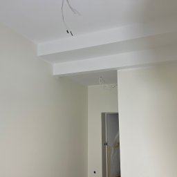 Roman Horditsa - Świeżo pomalowane na biało ściany i sufit z dekoracyjnymi belkami pod sufitem, widoczne wystające przewody elektryczne i zabezpieczone folią drzwi.
