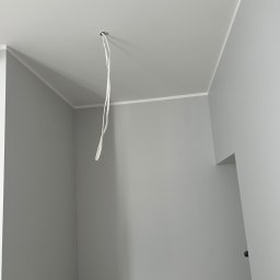 Roman Horditsa - Pomalowane na szaro wnętrze pokoju z wystającymi kablami ze stropu, widoczne listwy przysufitowe i otwór w ścianie.