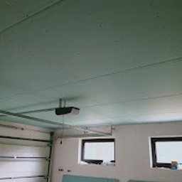 Roman Horditsa - Płyty KG na suficie garażu, widoczne okna, brama garażowa z automatyką. Surowy stan wykończenia, szare ściany, instalacja elektryczna.