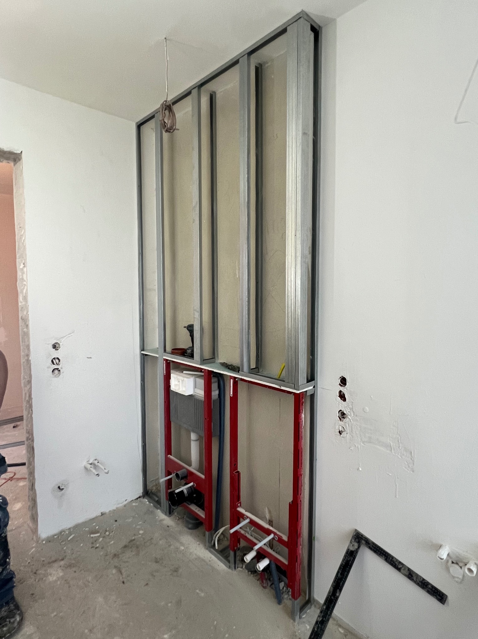 Stelaż metalowy pod zabudowę WC ze zbiornikiem, czerwone rury instalacyjne, widoczne przewody elektryczne w ścianie, w tle fragment niezabezpieczonego otworu drzwiowego, narzędzia na podłodze.