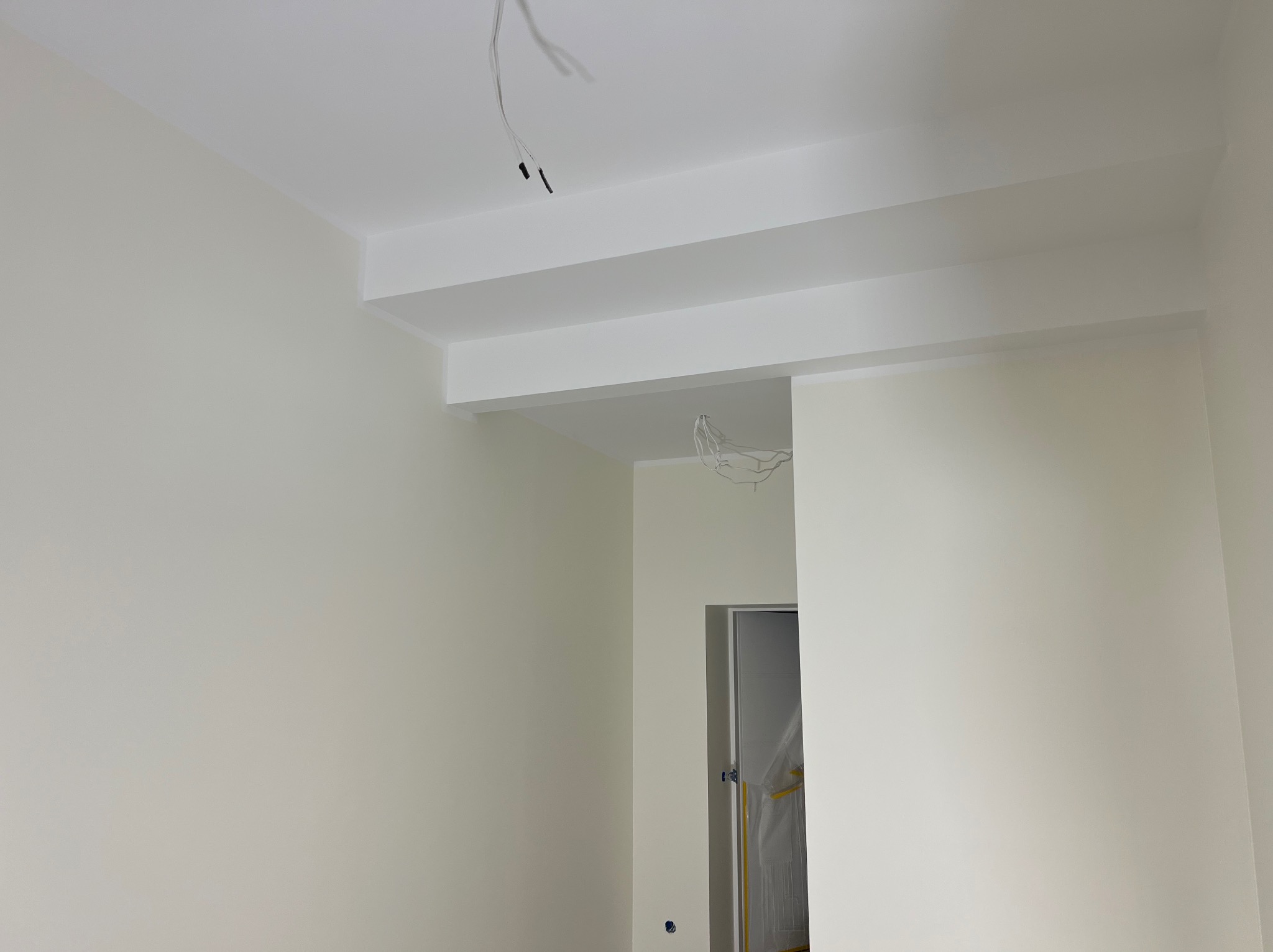 Świeżo pomalowane na biało ściany i sufit z dekoracyjnymi belkami pod sufitem, widoczne wystające przewody elektryczne i zabezpieczone folią drzwi.