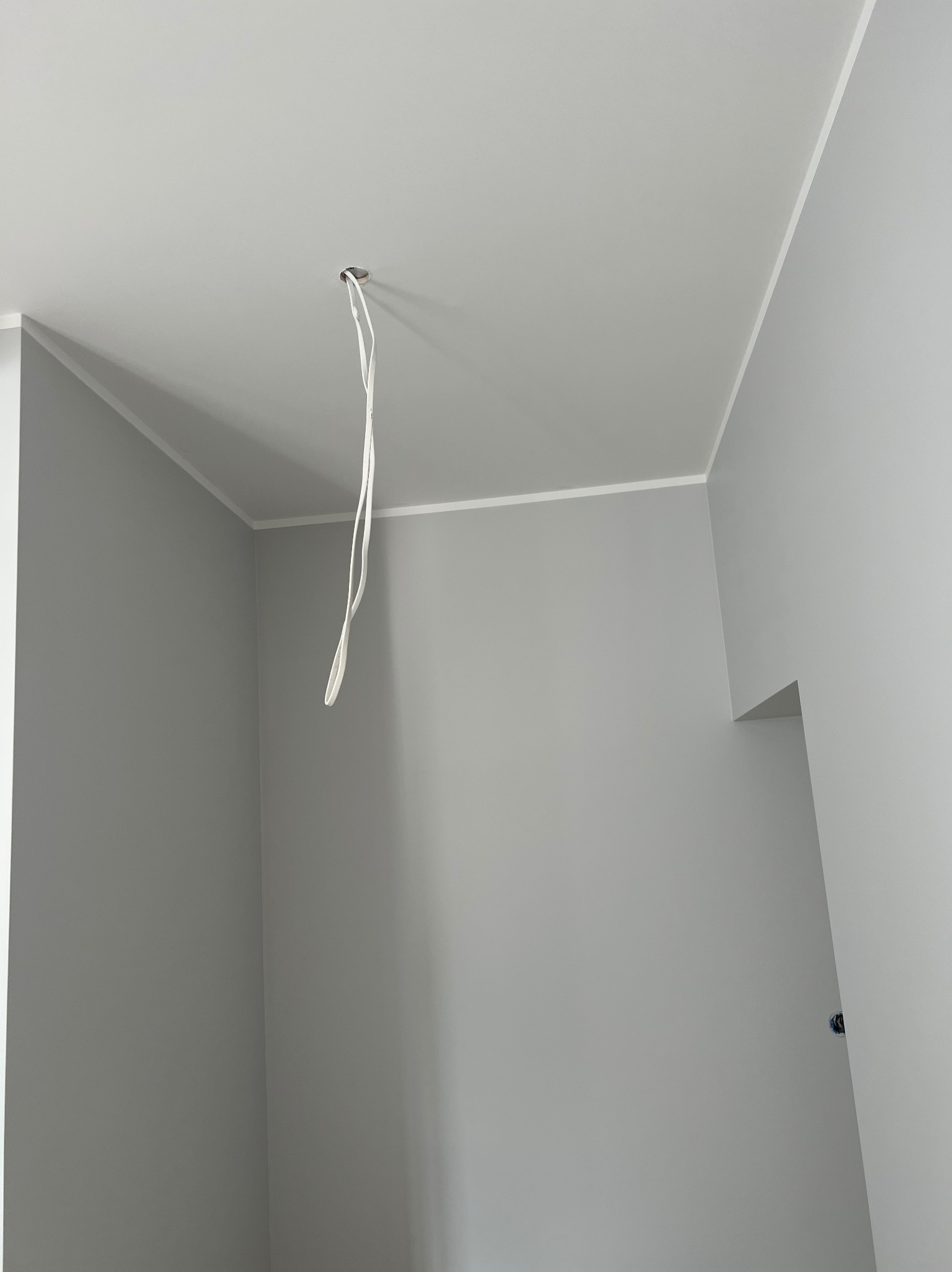 Pomalowane na szaro wnętrze pokoju z wystającymi kablami ze stropu, widoczne listwy przysufitowe i otwór w ścianie.