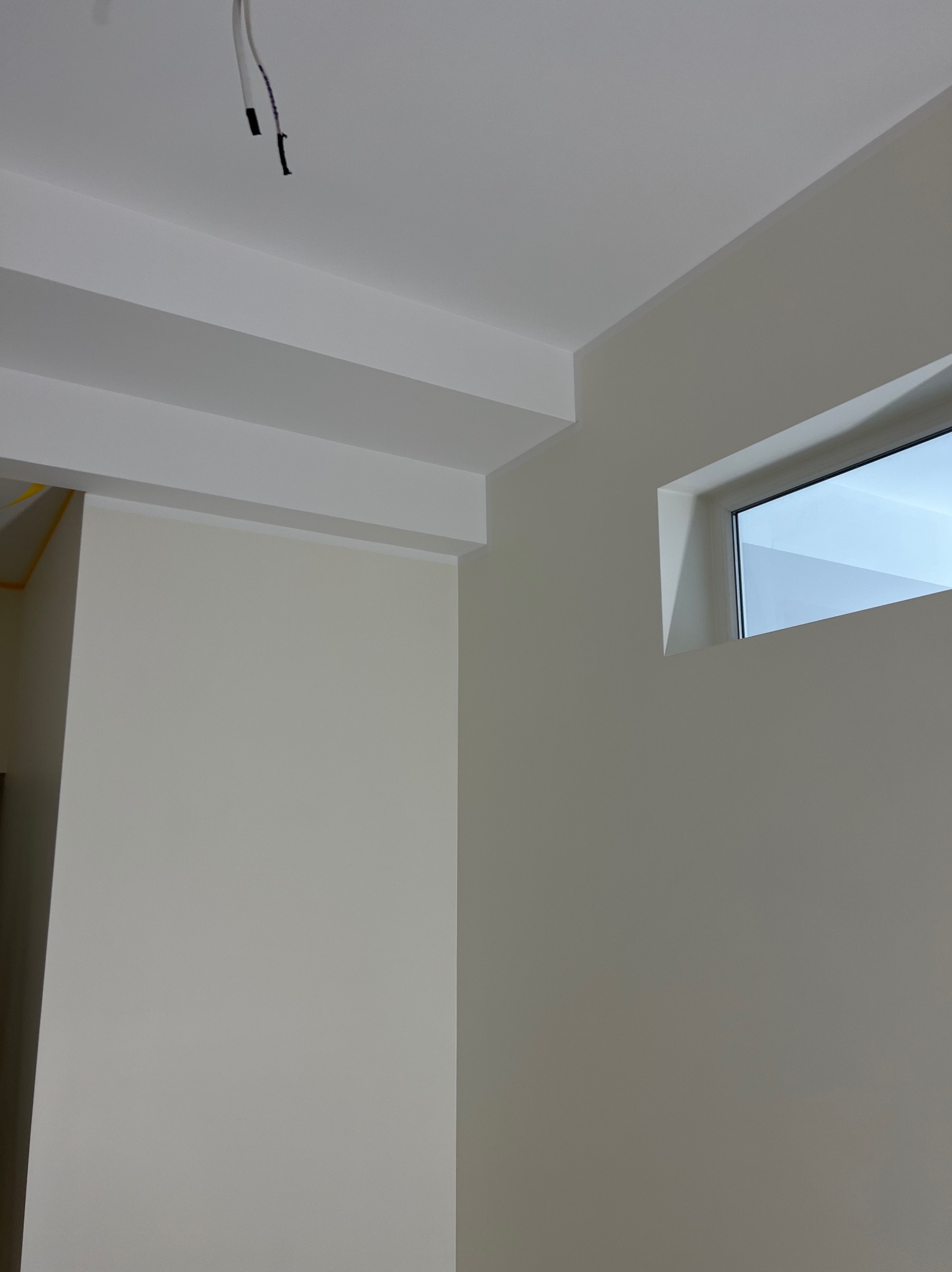 Wnętrze pokoju po malowaniu na jasny odcień beżu, widoczne fragmenty sufitu z listwami dekoracyjnymi, okno oraz wystające kable.