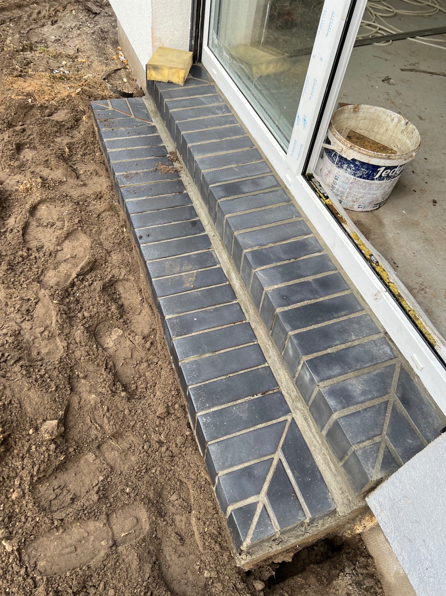 Wykończone schody wejściowe z ciemnej cegły klinkierowej, z widocznymi śladami butów w ziemi obok, żółta gąbka na parapecie i wiadro po farbie w tle.