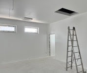 Białe wnętrze po gładziowaniu: gładkie ściany, sufit z otworem, okna i drabina. Minimalistyczny efekt wykończenia wnętrza, jasne oświetlenie.