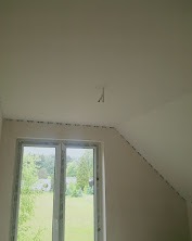 Wnętrze poddasza po gładziowaniu ścian i sufitu, widoczne okno z widokiem na zieleń. Minimalistyczne wykończenie, przygotowane do dalszych prac wykończeniowych.