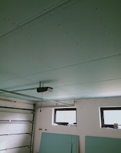 Płyty KG na suficie garażu, widoczne okna, brama garażowa z automatyką. Surowy stan wykończenia, szare ściany, instalacja elektryczna.
