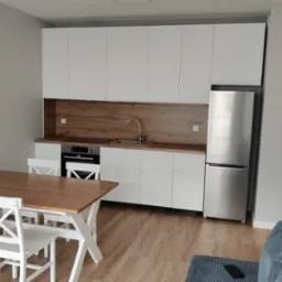 Nowoczesna kuchnia z białymi szafkami, drewnianym blatem i stołem z krzesłami. Uwagę przyciąga srebrna lodówka i drewnopodobny panel nad blatem.