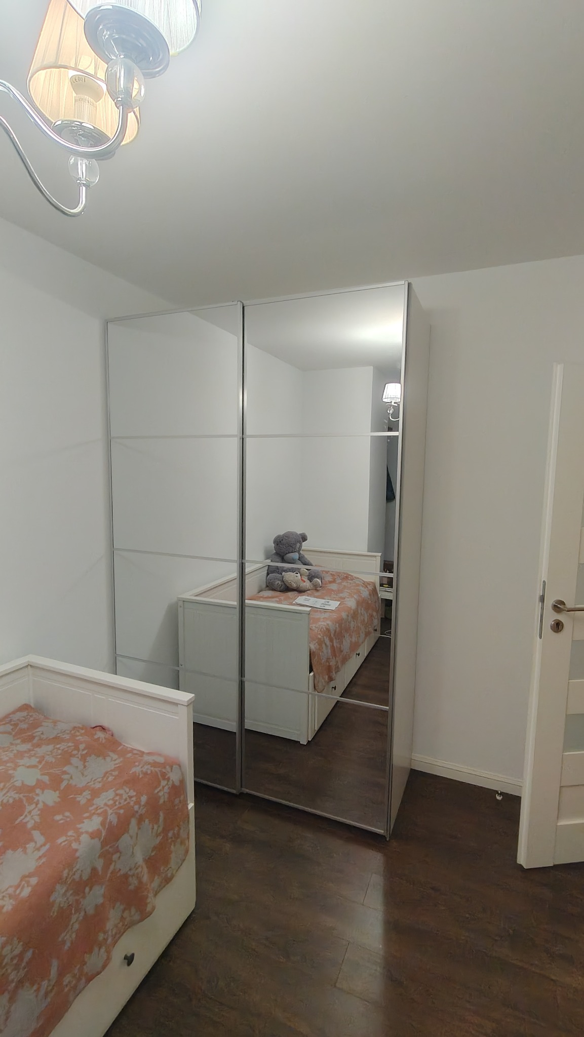 Biała szafa przesuwna z lustrami w pokoju dziecięcym, widoczne odbicie łóżka z pościelą w kwiaty i pluszowymi zabawkami.